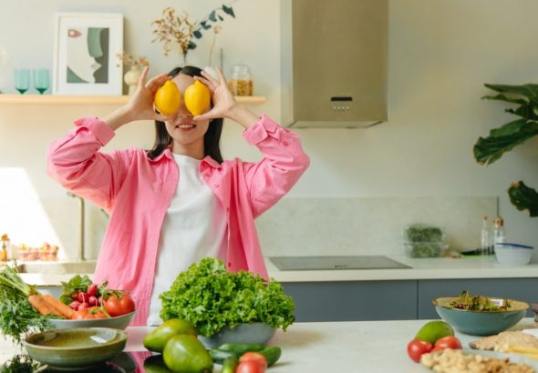 A woman playfully holding lemons over her eyes in a bright, lively kitchen setting filled with fresh vegetables.