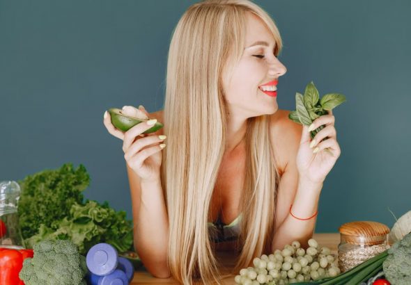 A smiling woman holding avocado and basil, surrounded by fresh vegetables and fruits for healthy eating.