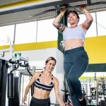 Two women practicing pull-ups in a modern indoor gym environment, emphasizing fitness and training.